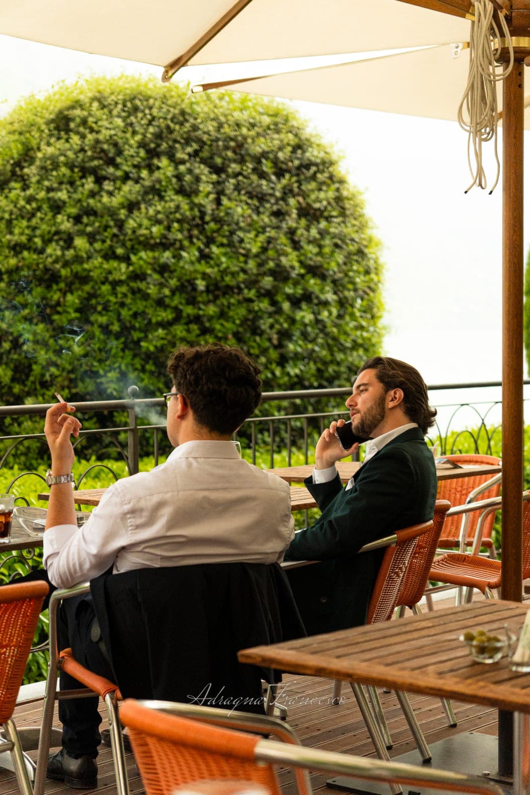 Two men talking and checking a phone at a table on a sunlit terrace